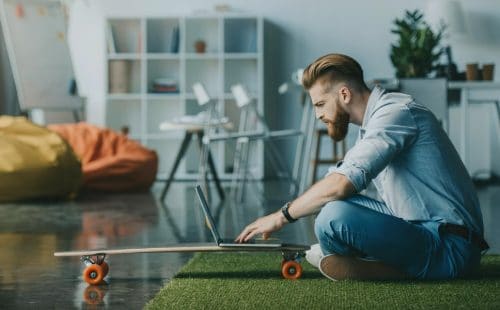 A man using a laptop perched on a skateboard in a light airy apartment. It looks like he is apply for a new job in Summer