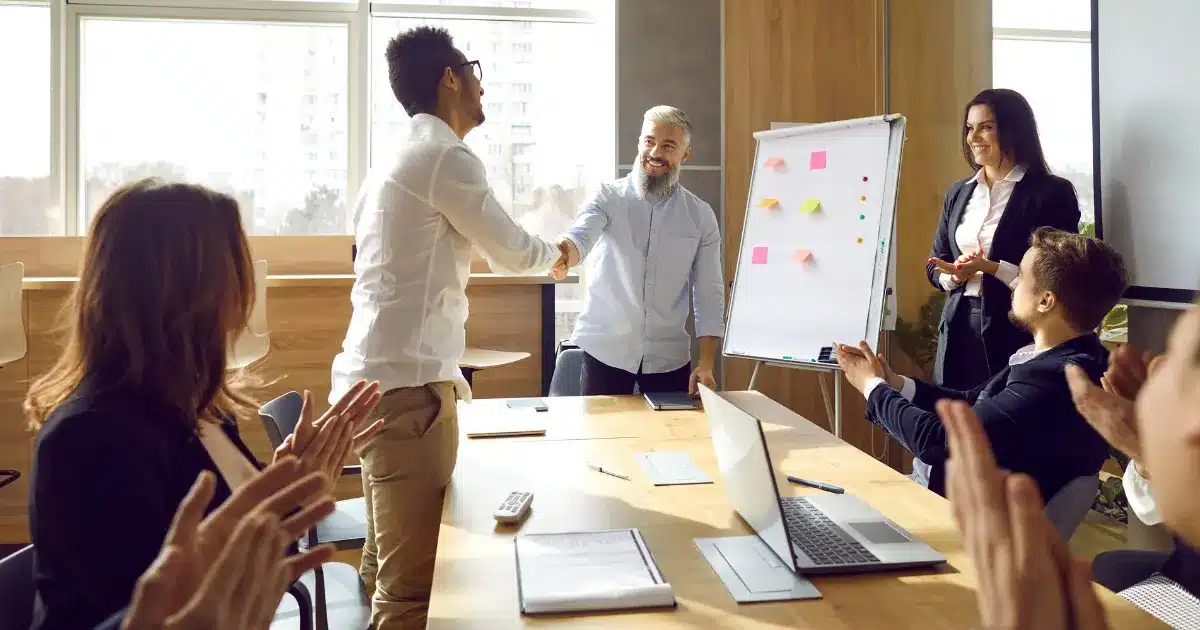 A manager congratulates a member of his team on his recent promotion in a meeting and other members of the smile and clap to give their approval