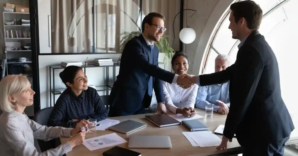 A hiring manager with glasses and a suit shakes hands with a new hire to welcome him into the team, while three women and a man look smiley