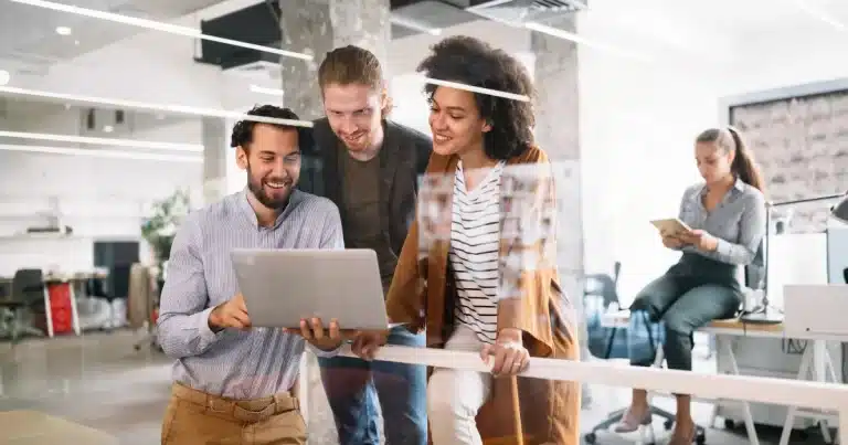 A team of tech workers look at their laptop and smile, while a woman sat on her desk in the background looks at her ipad