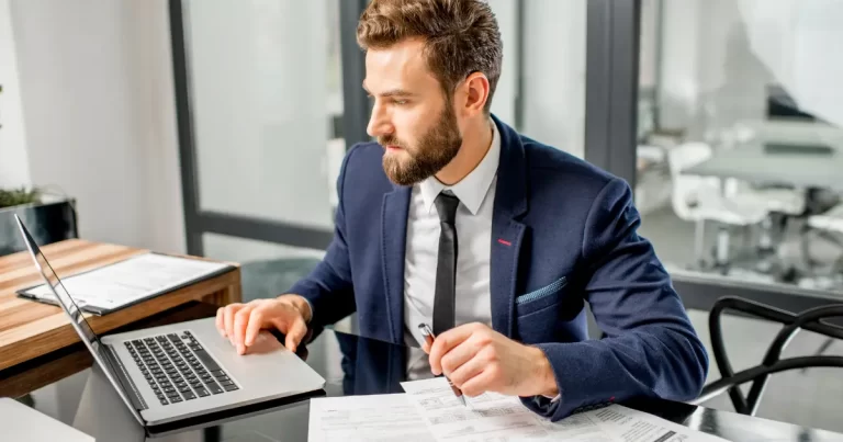 A male manager looks busy, juggling some important tasks, while looking at his laptop and some reports on his desk