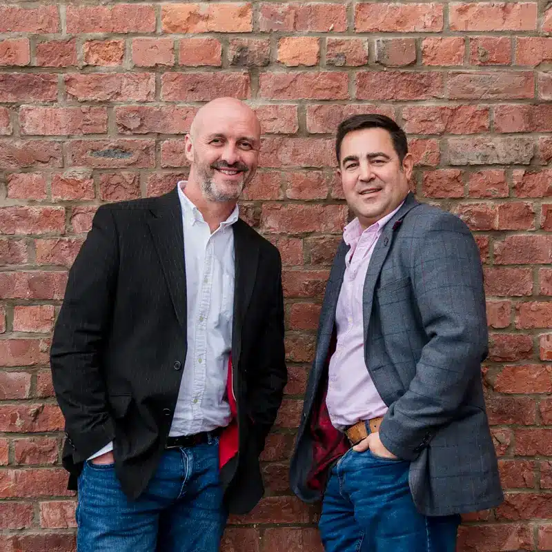 David Berwick and Nick Derham, directors of Adria Solutions, smile while posing against a bricked wall