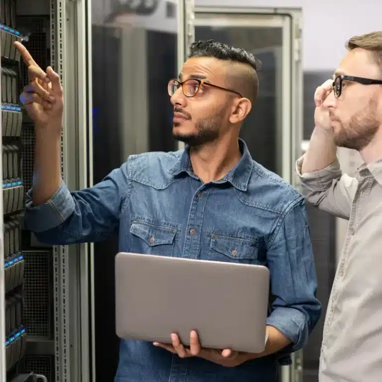 Two male IT network professionals, a junior Network Administrator and a more experienced Network Engineer, discuss a project