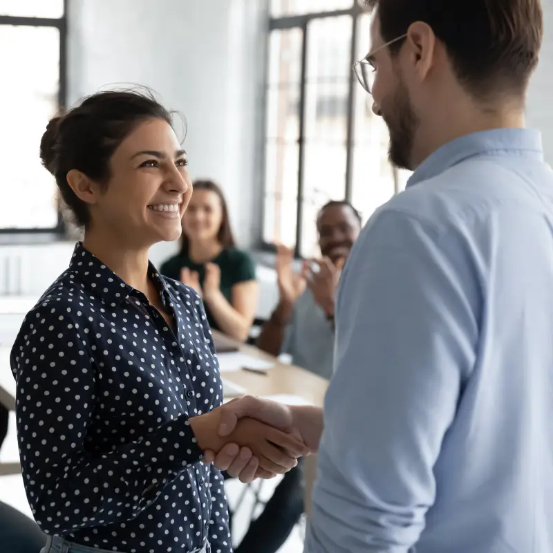 A woman who has just been promoted to team lead shakes the hand of her manager, while other team members aplaude the decision in the background