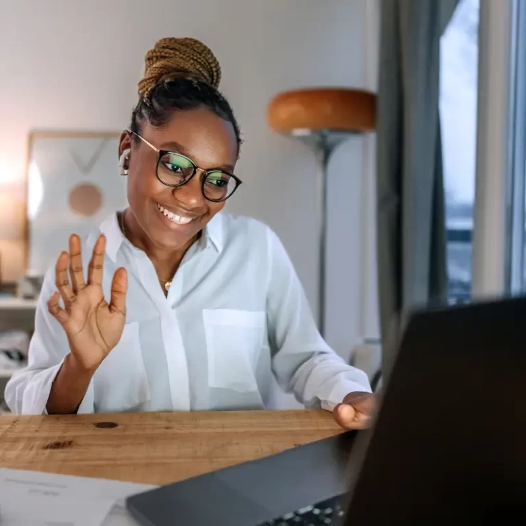 A black woman with glasses says hi to her colleagues, working remotely