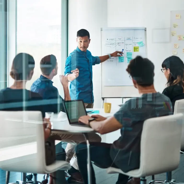 A team leader discusses ideas and the state of their projects and plans for 2025 with his team, using a board as a visual in a meeting room with four more colleagues.