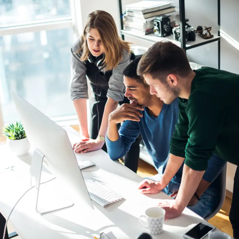 A female Digital Marketing Manager in Glasgow oversees a campaign with her marketing team, including two men, on the iMac of one of them.