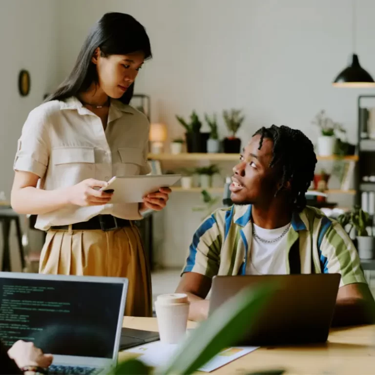 Two Gen Z workers, an Asian woman standing up with an iPad and a dark-skinned man sitting down with a laptop, chat in an office with good vibes