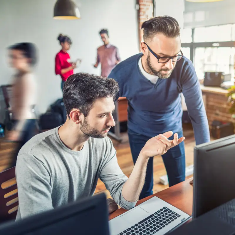 a senior software developer stands up, looking at the code a junior programmer wrote and overseeing his work