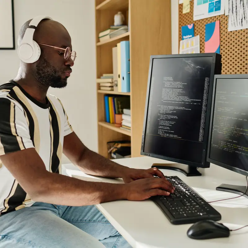 A black man in his mid 30s works on some code with headphones on and two screens